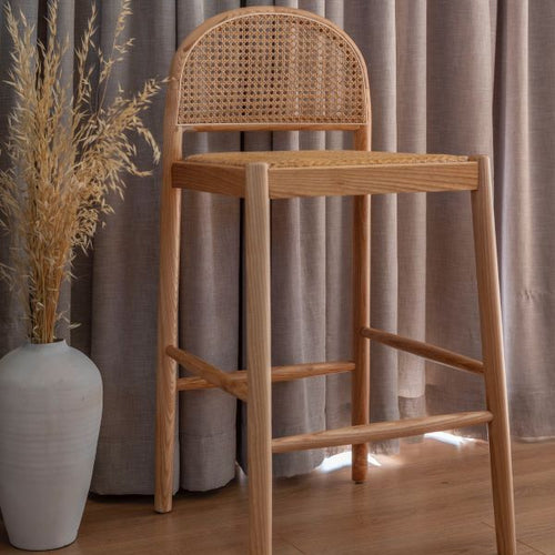 Wooden high chair with wicker backrest next to a white vase with dried plants against a curtain backdrop