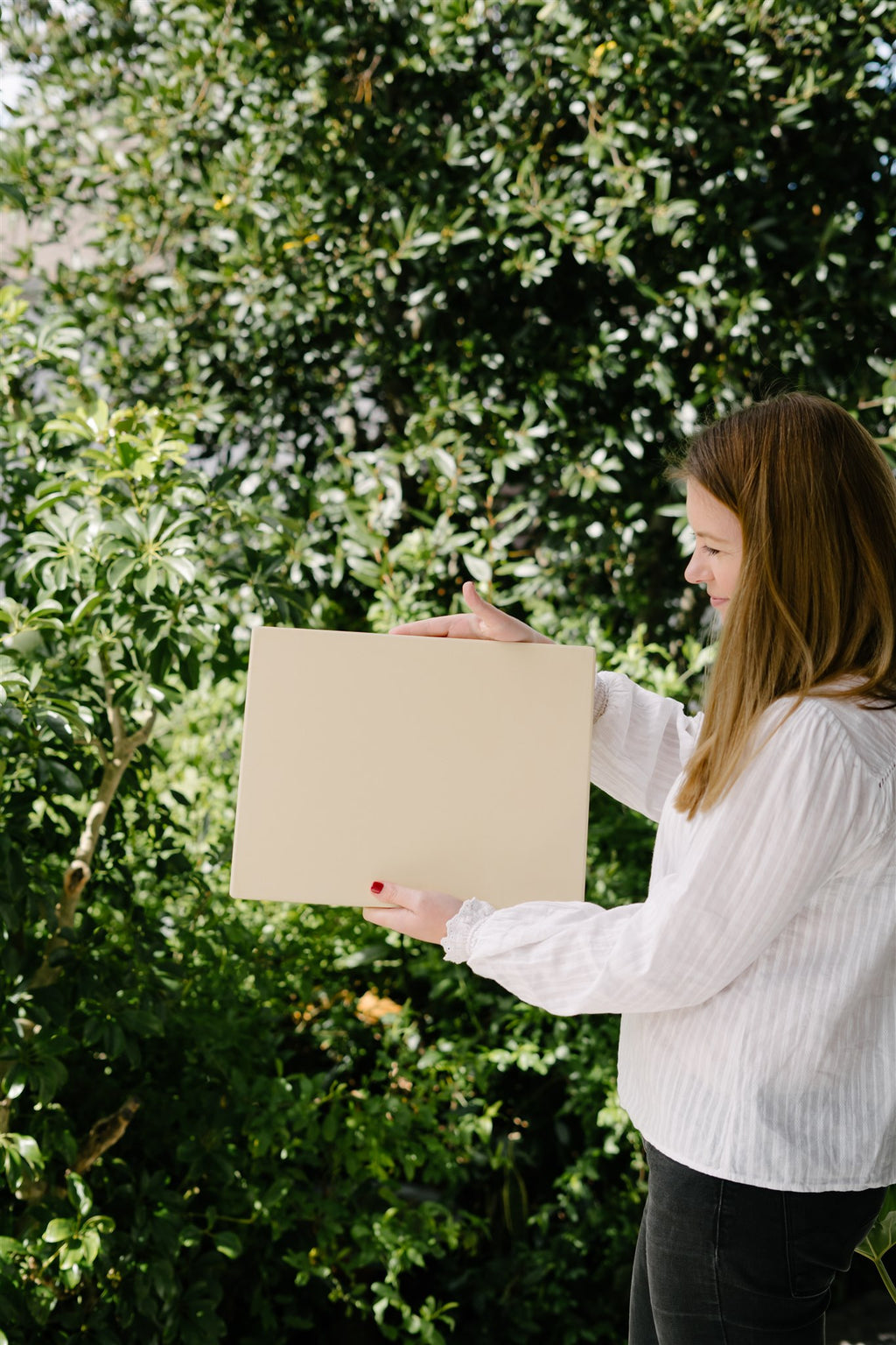Woman holding a beige square in front of a green leafy background