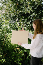 Woman holding a beige square in front of a green leafy background