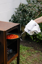 Person cooking outdoors using a metal grill with food on a pan, wearing a white glove.