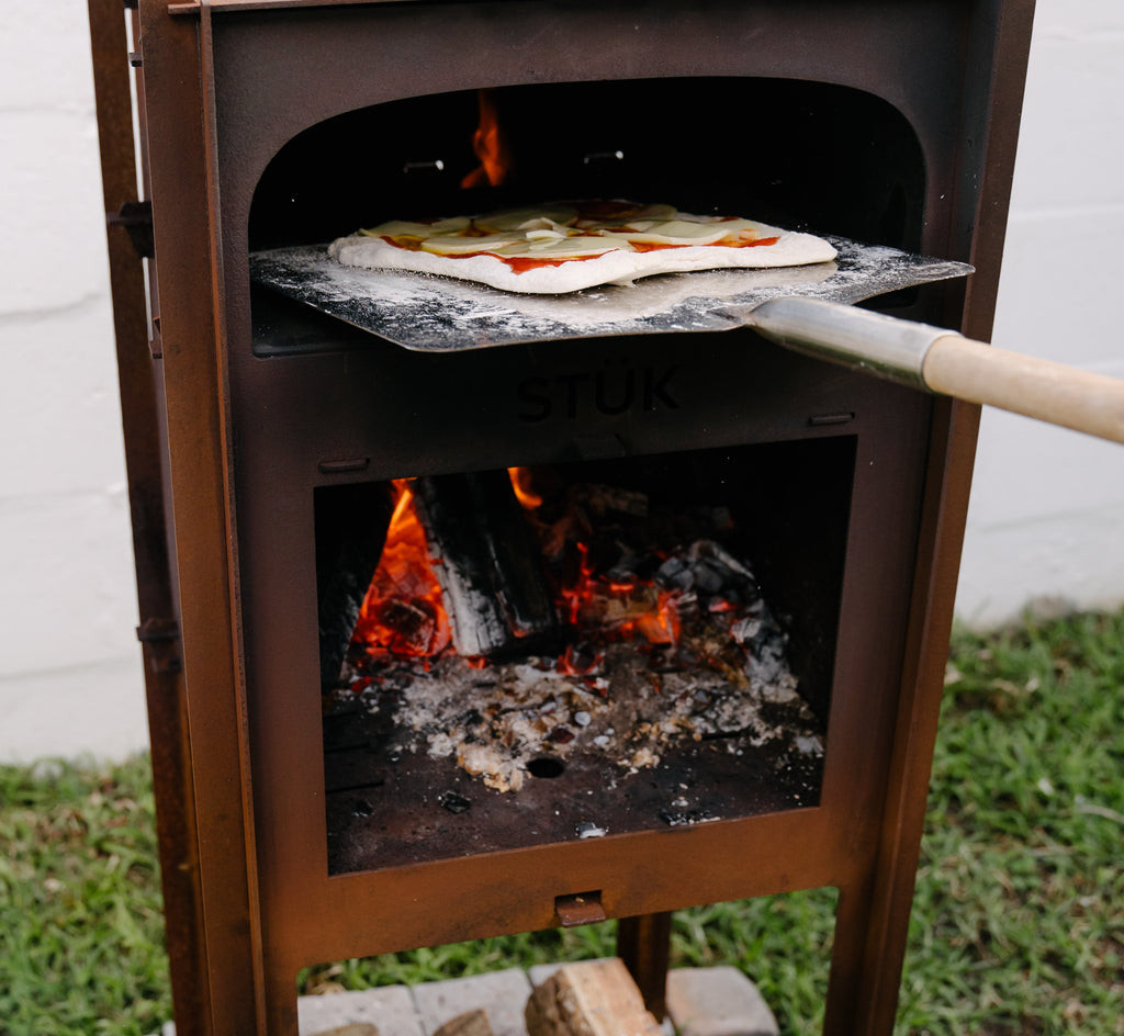 Wood-fired pizza oven with pizza being cooked and firewood on a brick base.
