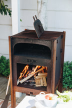 Outdoor wood-burning stove with flames and 'STÜk' branding, set against a white wall.