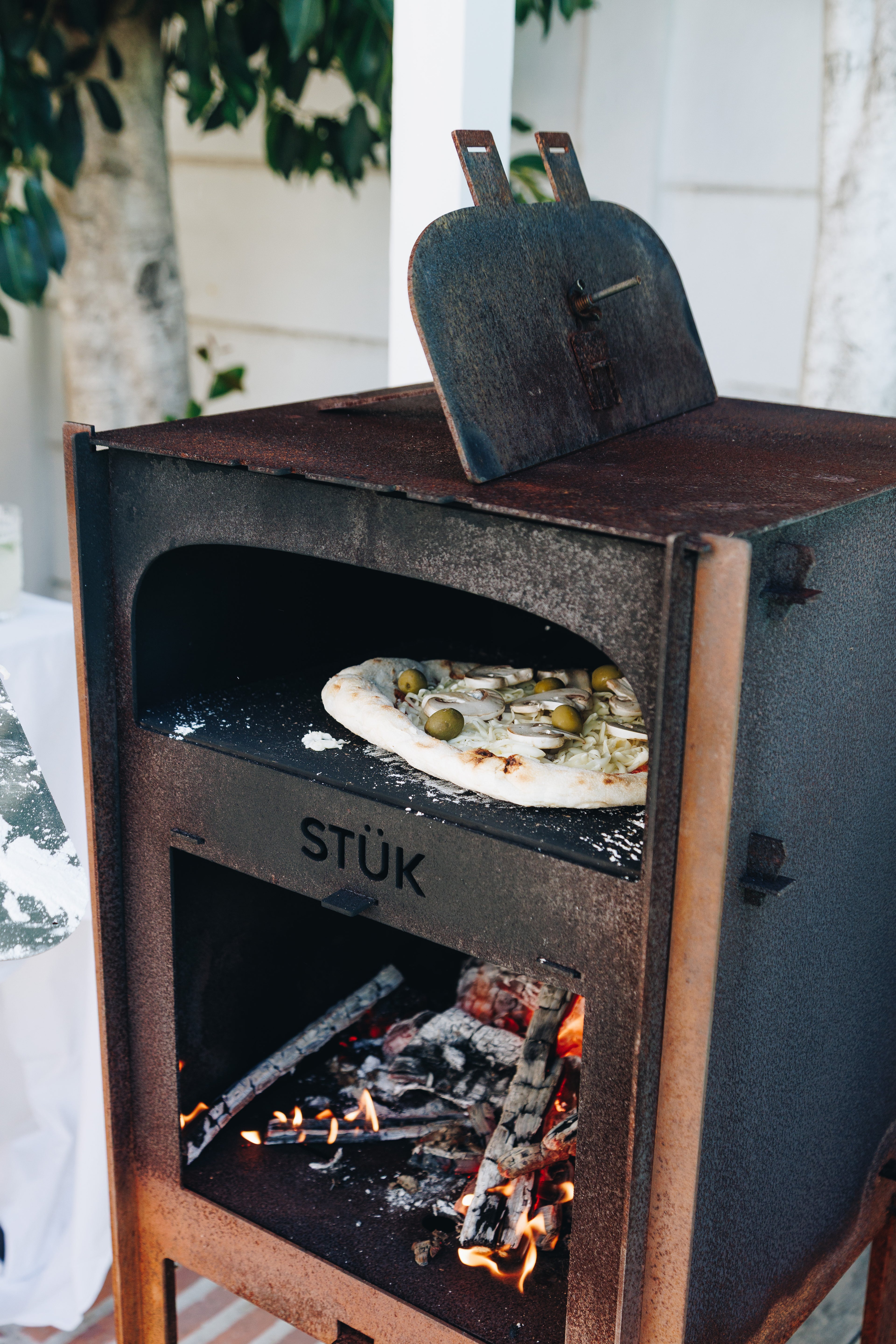 Wood-fired oven with a pizza inside, branded 'STÜk', outdoors.