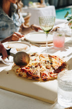 Person cutting a pizza on a wooden board with a glass of pink drink and a wine glass in the background.