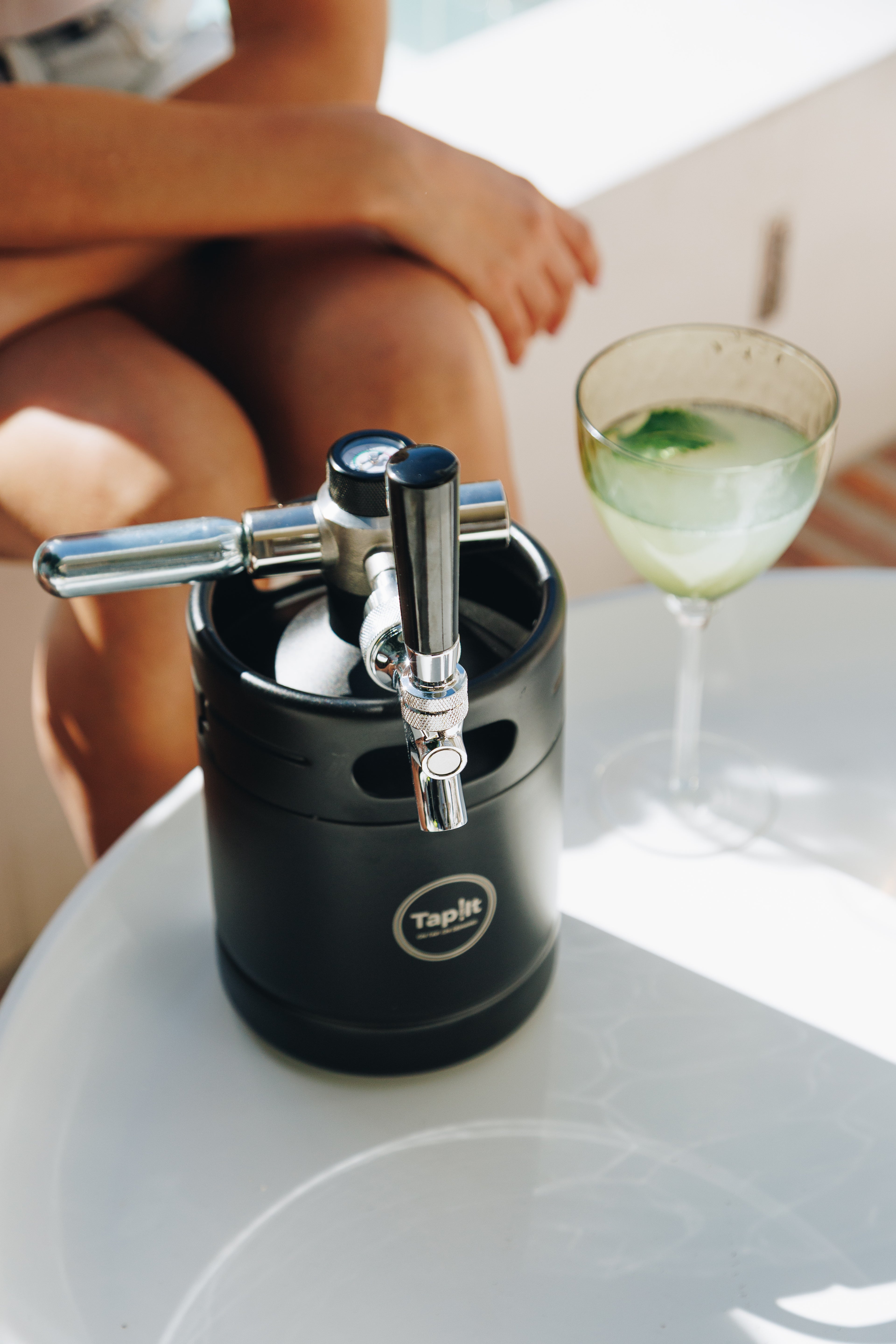 Black keg with a tap on a white surface next to a glass of green liquid.