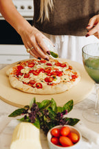Person preparing a pizza with tomatoes and herbs on a kitchen counter.