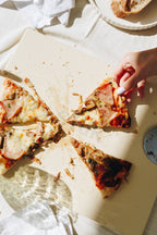 Person picking up a slice of pizza from a wooden board with other slices on a white surface.
