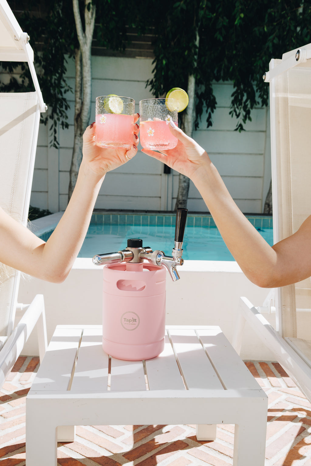 Two people holding pink drinks with lemon slices above a pink beer keg by a pool.