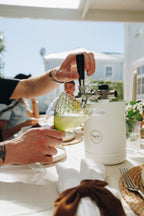 Person pouring a drink from a white pitcher labeled 'tapit' into a glass at an outdoor dining table.